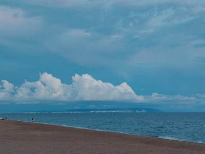 The image depicts a tranquil scene of a beach and the sea, under a cloudy sky. The foreground is dominated by a sandy beach, with the texture of the sand suggesting it is dry. In the middle ground, the turquoise sea stretches towards the horizon, with gentle waves lapping at the shore. A faint white line of foam marks the edge of the water on the sand.
Further out, a distant coastline is visible, marked by a line of buildings that appear to be part of a settlement, nestled against a backdrop of low hills. Above the coastline, large, puffy white clouds are scattered across the blue sky, some of them casting soft shadows on the water below. The overall impression is one of a peaceful day at the beach, possibly during the late afternoon or early evening, given the muted light.
The image's location is identified as Blagoveshchenskaya, Russia. This geographical context implies a coastal area within Russia.