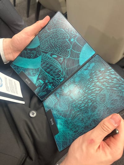 A person holds a black, foldable electronic device with a blue screen displaying drawings. The device is open, revealing two screens. The person is holding a stylus in their right hand and wearing a black suit jacket. A white card with printed text is visible near the bottom of the image. The location is implied to be somewhere in Huangdao District, China, though there is no direct visual confirmation of this. The focus of the image is the electronic device and the act of holding it.