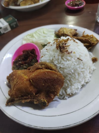 A close-up, overhead shot shows a plate of Indonesian food, likely in Kutalhoksukon, Indonesia, resting on a dark wooden table. The plate contains a generous portion of white rice, a piece of fried chicken, some fried shallots sprinkled on top of the rice, a small pink bowl of chili paste (sambal), and a few pieces of fried snacks. To the left of the plate, a portion of raw cabbage is visible. In the background, a blurred cigarette pack and a small pink dish with a dark substance are present. The lighting is dim, suggesting an indoor setting, possibly a restaurant or a home dining area.