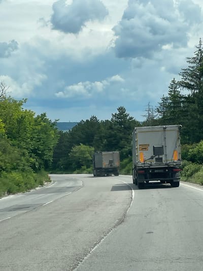 The image shows two box trucks driving on a curved asphalt road, possibly in a mountainous or hilly area. Lush green trees line the road on both sides. In the background, a sky with fluffy white clouds is visible. While the general location appears to be rural, the exact location in Osenets, Bulgaria cannot be determined from the image. There are no people visible in the image.