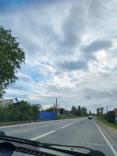 The image captures a view from inside a vehicle looking forward through the windshield onto a rural road in Malaya Bod'ya, Russia. The bottom portion of the image shows the vehicle's dashboard and the lower edge of the windshield, with several dark, circular smudges indicating reflections or dirt on the glass. The two-lane asphalt road extends into the distance, curving slightly to the right, with a solid white line delineating the right shoulder and a dashed white line separating traffic directions. Two passenger vehicles are visible ahead, traveling in the same direction; the lead car is dark-colored, followed by a lighter-colored vehicle.
On the left side of the road, a dense green tree partially obstructs the upper-left sky. Beneath the tree and alongside the road are various bushes and utility poles with overhead wires. A wooden building and a vibrant blue corrugated metal fence are visible, separating properties from the roadside. Further along the left, another house with a yellow exterior wall is partially visible. The right side of the road features a grassy shoulder, utility poles, and, in the distance, a house with a reddish roof. The sky overhead is predominantly cloudy with varying shades of grey and white, but patches of brighter blue sky are visible, particularly towards the left, indicating partial cloud cover. No people are explicitly visible in the scene.