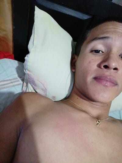 A light-skinned person lies on a bed, looking into the camera. The individual has short dark hair, dark eyes, and full lips. They wear a gold necklace with a small charm. A white pillow with a subtle floral pattern sits behind the head. The person's bare shoulder is visible. Bed sheets with a light blue and white pattern are partially visible. A dark headboard frames the scene.