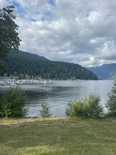 The image depicts a scenic view of a marina and coastline at Fisherman's Cove in the District of North Vancouver, Canada. In the foreground, a grassy area with some dirt patches leads down to the water's edge, with bushes framing the scene. The water itself is calm with ripples reflecting the cloudy sky. A marina filled with numerous sailboats is visible in the middle ground, lining the shore. Beyond the marina, a densely forested hillside rises, dotted with houses. In the distance, mountains form the backdrop, their peaks softened by the overcast weather. On the water in the middle ground, two people are visible on a small, inflatable raft, appearing to be paddling. The overall atmosphere is tranquil and natural, with a blend of human activity and the surrounding wilderness.