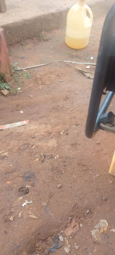 The image shows a close-up view of a dirt and concrete ground surface in Baissa, Nigeria. A large, translucent yellow plastic jug filled with a light yellow liquid is visible on the right side of the frame, placed on the concrete. The jug has a handle and a cap. To the left of the jug, a dark metal object, possibly part of a chair or other furniture, is also visible. The ground is composed of reddish-brown dirt with scattered stones and small patches of dry grass. Some debris, including a piece of white and blue striped material and some crumpled plastic, is present on the ground. The scene appears to be outdoors, under daylight, with no specific indication of time of day or weather conditions beyond the general lighting. The overall impression is of an everyday outdoor setting, possibly a yard or a roadside. There is no visible text in the image.