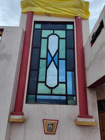 The image shows a building with a large stained-glass window. The window is framed by black mullions and features panes of blue, green, and white glass arranged in a geometric pattern. Two red cylindrical columns flank the window, resting on tiered bases. Above the window, a yellow tarp is draped over the top of the building. To the left of the window, a white angled wall extends outwards, and to the right, a portion of a brown wooden door is visible. A small framed image, possibly a religious icon, is mounted on the wall below the window. The building appears to be made of rough-textured plaster. The sky above is overcast. The image is taken from a slightly low angle, looking up at the building.