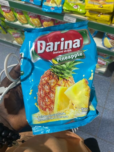 An eye-level shot captures a person holding a package of "Darina instant drink Pineapple" in what appears to be a store aisle in Goderich, Sierra Leone. The package is predominantly blue, featuring a large red oval logo with "Darina" in white, and text indicating "instant drink Pineapple." An image of a whole pineapple with cut slices and splashes of juice adorns the front. Below the English text, the French translation "Boisson Instantanée Aromatisée à l'Ananas" is visible.
The person's hand, dark-skinned, is holding the package, with a white cable (possibly from headphones or a charger) and a dark beaded bracelet or accessory visible near the wrist. In the blurred background, shelving units stocked with other colorful product packages, including yellow and green ones, suggest a supermarket or convenience store setting. The overall activity seems to be shopping or examining a product. Other partially visible text includes "Make," "Don," "rtion," "ns," "WINA," and "65."