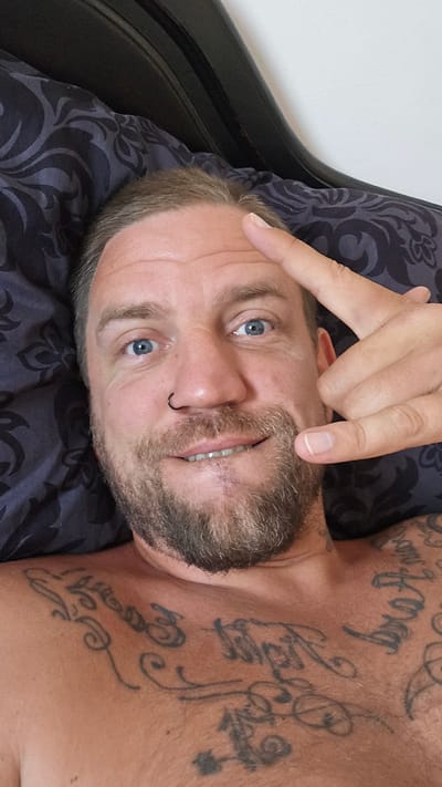 A man with fair skin and blue eyes is lying on his back on a patterned pillow. He has a short haircut and a salt and pepper beard. A small ring is visible in his left nostril. His right index finger is extended and pointing towards his forehead. Tattoos are visible on his upper chest and shoulder. The background includes what appears to be a headboard. No information indicates the location is Los Alcázares, Spain.