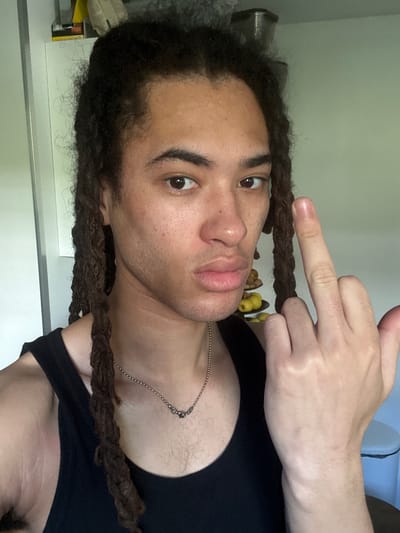 A young adult male with dark brown locs is centered in the frame, facing the camera with his head tilted to the left. He wears a black tank top and a silver necklace. His right hand is raised, extending his middle finger. The background is blurred, revealing a white cabinet surface, some fruit, and a blue object. No evidence indicates that the image was taken in Les Ulis, France.