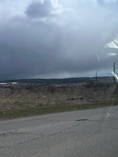 The image, taken from inside a vehicle, presents a wide rural landscape in Gol'yany, Russia, under a dramatic, overcast sky. The scene is dominated by a vast expanse of heavy, grey and white clouds, with a particularly dark, ominous band stretching across the mid-sky, suggesting a storm front or impending rain.
Below the distinct horizon, the land appears sparsely vegetated and dry, featuring brownish fields or uncultivated areas. A distant tree line or small forest can be seen. Multiple utility poles, carrying power lines, extend into the distance, paralleling a visible road. A small settlement or village with scattered buildings is discernible in the far distance, nestled within the landscape.
In the immediate foreground, the edge of an asphalt road with visible cracks and wear is present, bordered by dry grass and dirt. Reflections and glares on what appears to be a car window or windshield are prominent in the upper part of the image.
The overall atmosphere is rather subdued due to the heavy cloud cover. There is no discernible human activity or visible text in the image. The exact time of day is ambiguous, given the diffused light conditions.