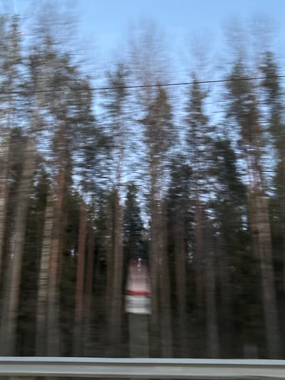 The image shows a view from a window, likely from a moving vehicle, perhaps a train, with blurred scenery including trees and what appears to be a building or train car passing by. The location is likely Yoksolovo, Russia. The blurry nature of the image indicates movement. There's a wire visible in the upper left area. The right side of the image reveals the edge of the window frame.