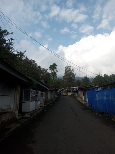The image shows a street scene in Mya Sit Kaing Ywar Thit, Myanmar. The road, made of dark asphalt, stretches into the distance between rows of buildings. The buildings are a mix of low, possibly one-story structures with varied construction materials and states of repair. Some have open fronts with window frames, while others are covered with blue tarpaulins. Debris is visible along the road and the edges of the buildings. Electrical wires crisscross the sky above. In the distance, a few vehicles are visible, and beyond them, a forested mountain landscape is partially obscured by clouds. The sky is a mix of blue and white clouds.