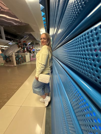 An individual with blonde hair and glasses stands leaning against a blue corrugated metal wall. They are wearing a light yellow long-sleeved shirt, light blue wide-leg jeans, and white sneakers. A small, white quilted purse with a chain strap is slung over their shoulder. The background suggests an indoor public space, possibly a mall or an arcade, with a tiled floor and various stalls or displays visible in the distance. The blue wall has a pattern of evenly spaced circular perforations. The scene is brightly lit. There is no indication this image was taken in Ermakovo, Russia.