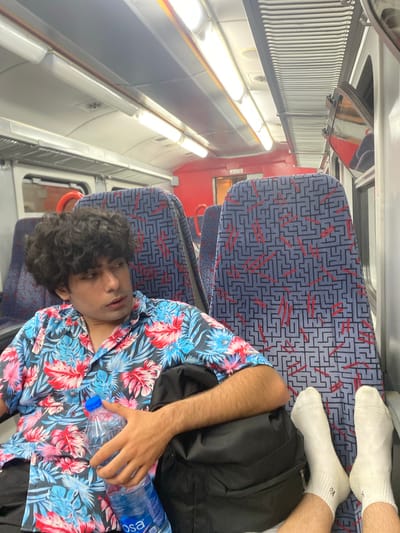 Inside a train carriage, a young man with curly dark hair lies sprawled across two seats. He is wearing a brightly patterned floral shirt, black trousers, and white socks. His feet are resting on one seat, while his head and torso lie across the other. A black backpack sits next to him, and he is holding a partially empty bottle of water. The train carriage has blue and red patterned seating and fluorescent lighting along the ceiling. The location is unspecified and not within Polje, Montenegro.