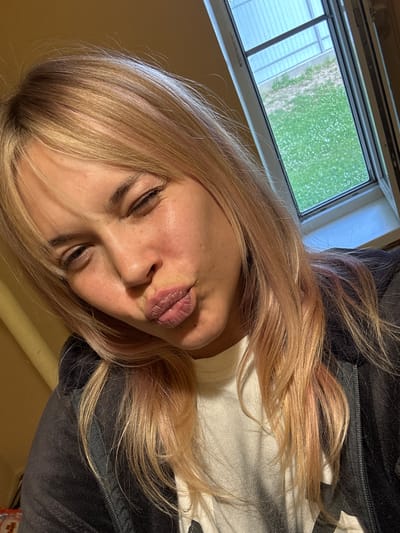 A young woman with blonde hair, tinged with pink, winks and puckers her lips at the camera. She is wearing a white t-shirt and a dark zippered hoodie. Behind her, a window reveals a glimpse of a grassy area with scattered white flowers, suggesting an outdoor setting. The wall to her left is a warm yellow. The scene is likely indoors, possibly a residential area in Naro-Fominsk, Russia, given the context provided.