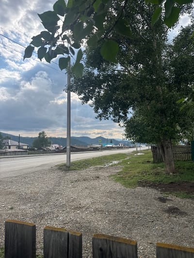 The image depicts a view of a road and buildings in a village, likely Yabogan, Russia, under a cloudy sky. In the foreground, a gravel shoulder is visible, bordered by a weathered wooden fence. A paved road with white lane markings runs horizontally across the midground. To the left of the road, a line of houses with gabled roofs and wooden fences can be seen. A tall utility pole stands near the center of the frame. On the right side of the road, a large tree with dense foliage shades the grassy area and a wooden fence. In the background, rolling hills covered in trees recede into the distance. A yellow triangular road sign with a black symbol is visible on the left side of the road in the distance. The overall scene suggests a rural or semi-rural environment. There are no people or animals visible in the image.