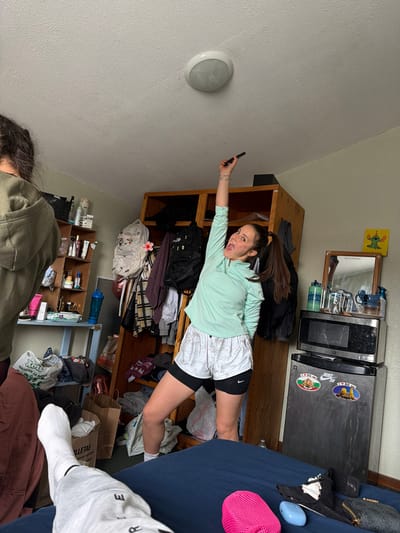 The image captures a cluttered indoor setting, likely a bedroom or dorm room, featuring two individuals. The primary subject, a young woman with long brown hair, is positioned dynamically in the mid-ground, appearing to "fly" or float horizontally. She wears a light green long-sleeved top and white athletic shorts over black compression shorts. Her right arm is extended, holding a black phone, while her left arm is bent. She has an open-mouthed, expressive, and playful look. Her legs are bent, and her left foot rests on a dark blue bed.

Behind her is a wooden shelving unit filled with items like black backpacks, folded clothes, and various personal belongings. To the right of this unit, a smaller wooden shelf holds a mirror, a blue mug, and a green water bottle bearing the text "DEINA." Above this shelf, a yellow drawing depicting a character and the phrase "WND IS THIS DIVA?" is affixed to the wall.

Further right, a microwave and a mini-fridge are stacked. The mini-fridge is adorned with stickers, including "NIKE SB" and two circular stickers prominently displaying "I ❤️ BG," likely referring to Bulgaria. Adjacent to the fridge, a dark blue bed is visible, covered with a blue blanket, a pink mesh hat, and a light blue oval object.

In the bottom left corner, the head and shoulder of a second person, wearing a dark green hoodie, are partially visible. At the bottom right, a foot in a white sock is seen resting on the bed. Additional objects scattered throughout the room include various shopping bags, a blue water bottle, and a cardboard organizer filled with toiletries. A white circular light fixture is mounted on the light gray wall in the top left. Visible text on other items includes "Rois," "Пекарни за ядки" (Bakery for nuts), "500940," "LLETAS," and "Materiales importados." The overall impression is one of a lived-in, active space.