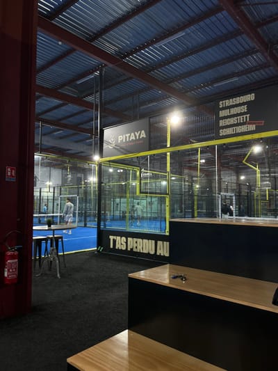The image shows an indoor padel court facility. Several courts enclosed in glass are visible, with players engaged in a match on one. Advertising banners for local cities and "PITAYA THAI STREET FOOD" are displayed overhead. The floor surrounding the courts is dark gray, and a counter with light wood trim and black paint stripes sits to the right. A red fire extinguisher is mounted on the lower part of the wall. The ceiling has metal corrugated panels.