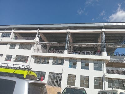 The image displays a modern, multi-story white building under a clear blue sky with sparse white clouds. The building's design features several floors, with the upper levels constructed as expansive open-air balconies or terraces. These open areas are supported by grey metal truss structures and are equipped with metal railings, suggesting they are public or commercial spaces.
Numerous people are visible on these upper balconies, appearing to be observing or participating in an activity, although the exact nature of the event is unclear. Several individuals are standing along the railings. The lower floors of the building are lined with multiple rectangular windows, many of which are open, indicating ventilation.
In the foreground, the lower section of the building is partially obscured by several vehicles. A prominent silver or grey SUV or van with a bright yellow cargo roof rack is visible on the left, alongside parts of other vehicles. A person in a red shirt is also seen on the ground level, near the building.
The bright daylight and clear sky suggest the photo was taken during the day, under favorable weather conditions. There is no discernible text or signage on the building or vehicles. The setting appears to be an active, possibly commercial or public gathering point, consistent with an urban environment in Siaya, Kenya.