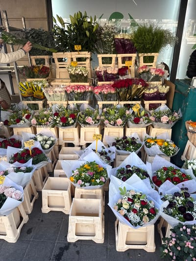 The image displays a densely stocked flower stall in Neuilly-sur-Seine, France, likely situated outdoors or at a market entrance. The floral display is structured using multiple tiers of light beige plastic crates.

The upper tiers feature large bunches of green foliage, including eucalyptus-like leaves, and tall green lily stalks (labeled "LYS 6€"). Numerous individual flower bunches, predominantly roses in white, pink, and red, are visible, wrapped in clear plastic sleeves, some with "MEIJER" branding. Small yellow sunflower-like arrangements are also present in the upper-left.

The lower tiers exhibit a profusion of round bouquets, each wrapped in white paper and tied with string. These bouquets contain diverse floral compositions, including red and pink roses, white gypsophila (baby's breath), yellow flowers, and dark burgundy ranunculus-like blooms. Several bouquets are marked with yellow price tags indicating "BOUQUET 30€." On the far right, bunches of white tulips and small potted pink plants are displayed.

On the far left, a person's forearm and hand, clad in a light-colored jacket, are visible reaching into a bundle of green foliage, suggesting an action of selection or arrangement. A dark, patterned handbag hangs from their arm. A segment of a dark jacket is visible on the far right, indicating the presence of another individual, possibly a vendor or customer. The background consists of a large, reflective glass surface, possibly a storefront window, showing indistinct reflections. The ground is a dark, paved surface.