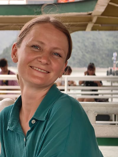 A woman with light skin and blue eyes smiles towards the camera. She is wearing a green button-down shirt. Behind her, other passengers are visible on what appears to be a boat or ferry. The background shows a body of water and tree-covered hills. A partial view of the roof structure of the boat is also visible. Given the location of Hải Phòng, Vietnam, the photo likely features Hạ Long Bay or a similar coastal location.