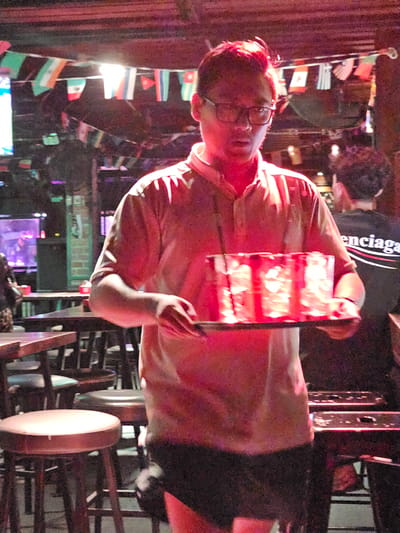 A man wearing glasses and a light-colored shirt is holding a red illuminated object with a black handle. He is positioned in what appears to be a dimly lit indoor venue, possibly a bar or restaurant, given the background details. The scene is filled with various objects and decorations, including flags from different countries strung up, shelves with items, and what looks like stage lighting or decorative lights in the background. The lighting is predominantly red and pink, casting a warm glow over the scene. The man's expression is difficult to discern clearly due to the lighting and image quality, but he appears to be engaged with the object he is holding. The overall atmosphere suggests a lively or festive environment. The background shows hints of a bar or club with tables and chairs, and what might be a television screen displaying purple lights. There is text visible on the back of the man's shirt which appears to read "enciaga" with a logo.