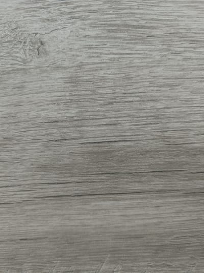 This is a close-up, abstract photograph of a light grey wooden surface, likely a table or a wall, with a visible wood grain pattern. The texture is characterized by horizontal lines of varying shades of grey, with darker lines indicating the grain and lighter areas suggesting the wood itself. There is a prominent knot or imperfection in the wood towards the upper left portion of the image, adding a natural element to the otherwise uniform pattern. The lighting appears even, with no distinct shadows, suggesting an indoor setting with diffused light. The image is a flat, two-dimensional representation of the material, focusing solely on its texture and color. There are no discernible subjects like people or objects, nor is there any indication of an activity or event. The location context provided is "Taraz city administration, Kazakhstan," which is not visually evident from the image itself. There is no visible text within the frame.