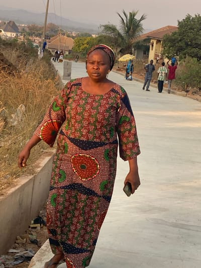 An African woman in a patterned dress stands on a concrete ledge. In the background, several people walk along a road near buildings and vegetation. The woman holds an object in her right hand. The landscape includes a power pole and distant hills. The scene is captured in a slightly tilted perspective, giving a dynamic feel to the composition. The lighting suggests it is early morning or late afternoon.