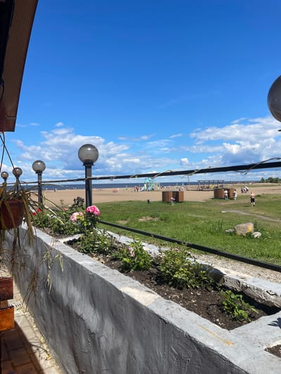 This image depicts a sunny day at a beach with a sandy shore and a body of water. In the foreground, there is a concrete planter box filled with green plants and some pink flowers. Behind the planter, a black metal railing runs horizontally. Several outdoor lamps with globe-shaped lights are visible along the railing. Beyond the railing, a beach area is populated with a few people, some children playing on playground equipment, and a few small wooden structures. The water appears calm, and the sky is a bright blue with scattered white clouds. The scene is likely a public recreational area. The location is the beach in Belozersk, Russia.
