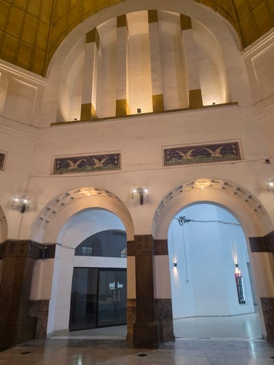 This media file captures an interior view of a building with architectural details suggesting a historical or culturally significant location in Medan, Indonesia. The scene is characterized by arched openings, decorative tiles featuring birds, and textured ceilings.
The main subjects are the architectural elements themselves. We see large, rounded arches framing doorways or passages. One arch reveals a brightly lit interior with a white wall and a digital display board. Another arch leads to a glass-door entrance with handles. The walls are adorned with rectangular decorative tiles, each depicting two stylized birds in flight against a patterned background. The ceiling in the foreground is covered with a textured golden material, possibly metal or painted plaster, with a repeating geometric pattern.
The lighting suggests it is likely daytime, or at least the interior is well-lit, creating a bright and somewhat cool atmosphere, especially in the areas visible through the arches. The overall mood is one of calm and possibly historical reverence. There are no people visible in the scene, nor any indication of a specific activity or event.
Notable details include the contrast between the white architectural elements and the darker, possibly stone, structural supports. The digital display board visible through one arch shows text that is partially legible but appears to be informational, possibly indicating train or bus schedules, given the context of Medan as a transportation hub. There is also a sign on the glass doors that is too small to read. Small dark specks are scattered across the walls and surfaces, possibly dust or debris. The floor is tiled with a marble-like pattern. The presence of visible electrical wires suggests ongoing maintenance or the presence of modern infrastructure within the older structure.
