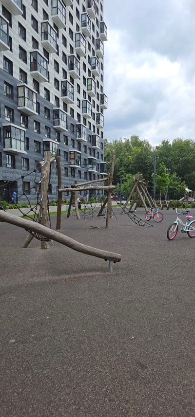 The image shows an outdoor children's playground in front of a modern residential building in Butovo, Russia. The building is tall with a dark gray and white facade, featuring many balconies and windows. The playground area has a dark, rubberized surface. Prominent features of the playground include wooden climbing structures with netting, a fallen log that appears to be part of the play equipment, and two children's bicycles, one light blue and white with pink accents, and another pink bicycle. A person in a blue garment is visible walking on a paved path to the left of the playground, near the building's entrance. The background consists of a dense line of green trees and a cloudy sky.