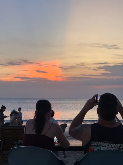 The image captures a sunset scene on a beach in Kuta Selatan, Indonesia. In the foreground, two individuals are seated at tables, their backs to the camera, facing the ocean. One person is holding up a phone to capture the sunset. To the left, a group of people are visible on the beach, including a woman in a hat and a child. The ocean is calm, with gentle waves lapping the shore. The sky is a canvas of warm colors, with hues of orange, pink, and purple dominating the sunset. The atmosphere is serene and relaxed, suggesting a leisurely evening on the beach.