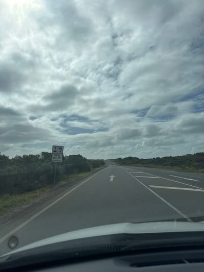 A highway stretches into the distance under a cloudy sky. An arrow is painted on the road, indicating the direction of travel. A sign on the side of the road reads "Drive on Left in Australia." The road is bordered by trees and vegetation. The view is from inside a vehicle.
