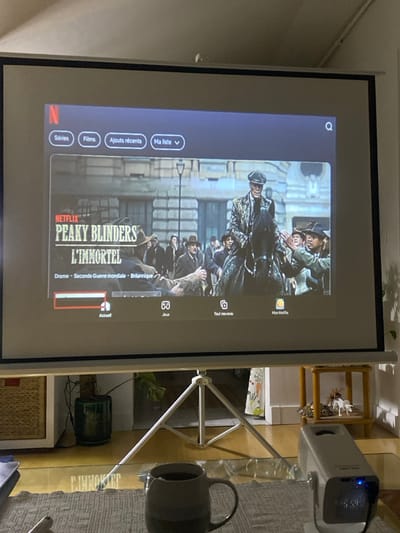 A digital image captures an indoor scene within a room in Boulogne-Billancourt, France, centered on a projected Netflix display. A white projector screen, supported by a three-legged white tripod stand, occupies the central frame. The screen displays the Netflix interface in French, featuring the title "Peaky Blinders L'IMMORTEL," categorized as "Drame - Seconde Guerre mondiale - Britannique." A red playback bar is visible near the bottom left of the projected content. The top navigation bar shows "Séries," "Films," "Ajouts récents," "Ma liste," and a search icon. Below the main content, icons for "Accueil," "Jeux," "Tout nouveau," and "Mon Netflix" are visible.
In the foreground, a dark grey mug with a tea bag string protrudes from it, resting on a light grey textured placemat on a transparent glass table. A portable, light-colored projector with a glowing blue indicator light is positioned on the right side of the glass table. A partial view of an electronic device, possibly a laptop or tablet, is visible on the far left of the table.
The background reveals a domestic environment. To the left of the tripod stand, a light brown wicker basket sits on the floor next to a dark green potted plant. To the right, a wooden two-tiered shelf unit holds white decorative figurines. A white wall, light-colored flooring, and a white sloped ceiling are visible. A patterned curtain is partially visible behind the tripod stand on the right.