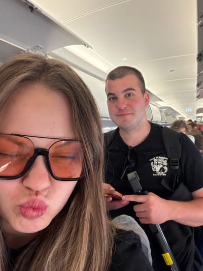 The image depicts two individuals on an airplane. The person in the foreground, on the left, is a female with long, brown hair, wearing large, orange-tinted sunglasses and making a puckering gesture with her lips. She is partially obscuring the face of the person behind her. The second individual, on the right, is a male with short, dark hair, wearing a black t-shirt with a graphic of a dog character and Japanese text that reads "TOO KAWAII TO DIE" and "かわいすぎて死ぬ". He is holding a phone and a small black device with a handle, possibly a camera or gimbal. The background shows the interior of an airplane cabin, with overhead compartments and other passengers visible further down the aisle. The scene is brightly lit, suggesting it is daytime. This image was taken in Orio al Serio, Italy.