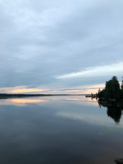 The image depicts a serene lake scene at dusk. The sky is filled with soft, grey clouds, with a faint band of warm, peachy light on the horizon, indicating the setting sun. The lake's surface is calm and reflects the sky and the distant treeline, creating a mirror-like effect. On the right side of the image, a cluster of dark green trees is visible, casting reflections on the water. The overall impression is one of tranquility and natural beauty. The location is identified as Svirstroy, Russia.