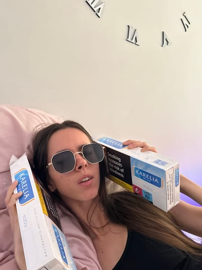 The scene depicts a female individual, appearing to be an adult, reclining on a light pink pillow or bedding in an indoor setting. She has long, dark brown hair and is wearing polygonal-shaped sunglasses with dark lenses and a gold-colored frame, along with a dark-colored top. Her mouth is slightly ajar. She is holding two large cartons of "KARELIA BLUE 100's" filter cigarettes, one on each side of her head. The carton on the viewer's right prominently displays the UK health warning "Smoking increases the risk of blindness" and a graphical warning of a pregnant woman with a crossed-out symbol. The carton on the viewer's left is partially visible, also showing "KARELIA BLUE 100's filter cigarettes." The background consists of a plain white wall, on which several black Roman numeral clock markers are affixed, specifically "XII," "II," and "IV." A subtle purple ambient light emanates from the right side of the wall. The specific location within Corby, United Kingdom, cannot be determined from the visual evidence provided; however, the cigarette packaging confirms a UK context.