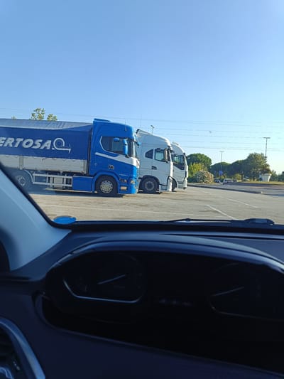 The media file shows a daytime scene in Riccione, Italy, from the perspective of someone inside a car. In the foreground, the dashboard of the car is visible, with a speedometer and other gauges. Beyond the car, a parking lot is occupied by several large trucks. One truck on the left is blue and white, with a large blue tarp covering its trailer. The tarp has white text that appears to read "FERTOSA." To the right of the blue truck are two white trucks, one parked slightly behind the other.
The parking lot is paved and marked with white lines. In the background, beyond the parking lot, there is a road with a few cars. Further back, there are trees and some greenery, suggesting an outdoor location. The sky is clear and blue, indicating fair weather. The sun is high in the sky, suggesting it is likely midday or early afternoon. There are no people visible in the media file. The overall scene depicts a typical roadside or service area with parked trucks under a bright, clear sky.