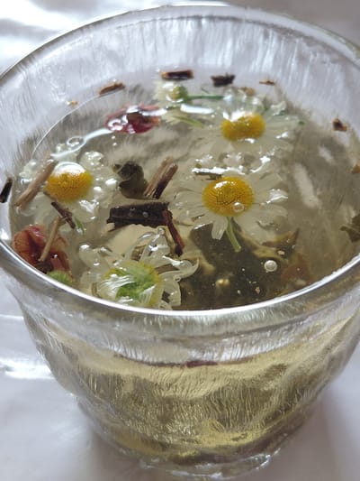 This is a close-up, top-down shot of a glass mug filled with clear liquid and visible herbal ingredients. The mug appears to be made of textured glass, with a ridged exterior. Inside the liquid, several small, white flowers with yellow centers, resembling chamomile, are floating. There are also small pieces of what appear to be dried plant matter and dark brown fragments, possibly cinnamon sticks or other dried herbs, dispersed throughout the liquid. Bubbles are visible on the surface of the liquid and attached to some of the floating ingredients. The overall impression is of a brewed herbal tea. The background is a soft, white surface, suggesting a clean or well-lit environment. There is no indication of the location being in Ертіс ауылдық округі, Kazakhstan; the image shows only the contents of the mug and the mug itself.