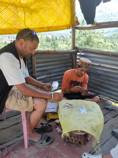 Two men are playing cards in an open-air structure made of corrugated metal and wooden planks in Kulang, India. The man on the left, wearing a white t-shirt, black vest, and khaki shorts, is sitting on a red stool and holding a fan of playing cards. He is wearing sunglasses on his head and a silver bracelet on his left wrist. The man on the right, wearing an orange t-shirt with "PUMA" printed on it and black shorts, is seated on the ground, wearing a traditional patterned cap. He is focused on a hand of cards. A large, yellow, burlap-like bag is spread on the wooden surface between them, serving as a makeshift table. Several playing cards are visible on the bag, including a four of clubs. The background reveals a mountainous landscape with scattered buildings and dense green trees, indicative of a rural or semi-rural setting. The structure appears weathered, with some fraying and damage to the materials.