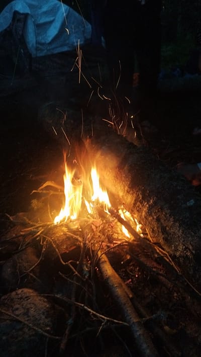 The image depicts a campfire at night, likely in a rural or wilderness setting, possibly near Tarasovskoe, Russia. The fire is fueled by dry branches and logs, with flames prominently displaying vibrant orange and yellow hues. Sparks are seen ascending from the fire, indicating active burning. In the background, a large fallen log acts as a backdrop to the fire, and a pale blue tent is partially visible, suggesting a camping scenario. The overall environment is dark, with the fire serving as the primary light source, casting a warm glow on the immediate surroundings. No people are clearly visible in the image, though their presence is implied by the campsite setup. The ground appears to be covered with dirt, rocks, and more branches.