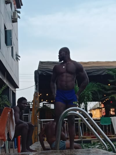 Three shirtless, dark-skinned men are positioned near a swimming pool. One is standing, wearing blue shorts, displaying a muscular physique. Another man sits on a chair with a towel draped nearby. A third man appears to be either sitting or lying at the pool's edge. Poolside rails are visible in the foreground. A building, a dark tent or canopy, and some greenery, including a palm tree, are visible in the background. The sky is overcast.