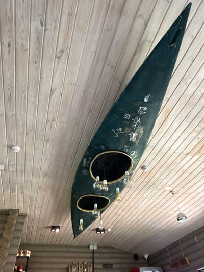 This is a low-angle shot of the interior of a building with a wooden plank ceiling and wooden walls. Suspended from the ceiling is a dark green canoe, decorated with white pictograms. The canoe has two oval cutouts, through which various light fixtures hang. Some of these are Edison-style bulbs, while others are more modern pendant lights. Several smoke detectors are visible on the ceiling. To the left of the frame, a rough-hewn wooden pillar extends upwards. The building appears to be a cafe or restaurant, indicated by the display shelves and menu boards visible on the lower wall. Based on the context of the canoe as decor, this is likely a themed establishment. The location is identified as Kupchegen, Russia. There are no people visible in the image.