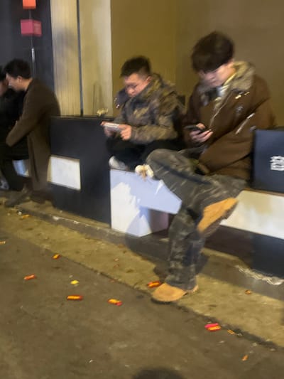 This media file captures three young men in an urban setting at night, located in 天宫殿街道, China.
In the foreground, one man is seen from behind, wearing a dark jacket. In the mid-ground, another man wears a camouflage-patterned jacket and is engrossed in his smartphone. Behind him, a third man, wearing a brown jacket with a fur-lined collar, also looks down at his phone. A black bag with an indistinct white logo rests nearby.
The scene appears to be an outdoor area, possibly a street or public space, illuminated by artificial light. The ground is dark asphalt or concrete, and scattered across it are numerous small, rectangular pieces of red and yellow debris, strongly resembling remnants of firecrackers. A white rectangular object, possibly a low stool or box, is situated between two of the men, and a dark surface, possibly a table or counter, is to their left.
The activity primarily involves the men checking their phones. The jackets suggest it might be a cool evening. The presence of firecracker debris indicates a recent celebration or event commonly observed in China. The overall mood is casual, with individuals focused on their devices.