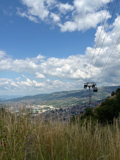 The image depicts a scenic view of a city nestled within a valley, as seen from a hillside covered in tall, dry grass. A cable car is prominently featured, suspended on wires that stretch across the frame, with a station and a gondola visible against the backdrop of the sprawling cityscape. The city appears to be situated on gentle slopes, with buildings and infrastructure extending as far as the eye can see.
The sky is a vibrant blue, dotted with fluffy white clouds, suggesting a clear and pleasant day. The time of day appears to be midday or early afternoon, based on the brightness of the sunlight and the shadows cast. The vegetation in the foreground, characterized by dry, golden grass, indicates a warm season, likely late summer or early autumn. The presence of the cable car suggests a tourist attraction or a mode of transport to an elevated viewpoint overlooking the city of Sarajevo. The overall atmosphere is one of natural beauty combined with urban development, offering a picturesque panorama. There is no visible text in the image.