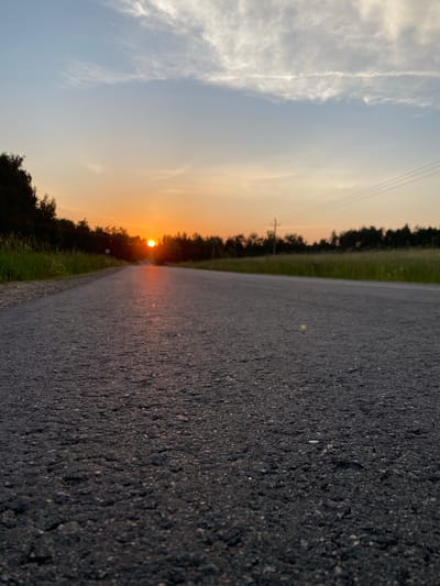 The image depicts a low-angle view of an asphalt road with a sunset in the background. The road takes up the right side of the frame, showing its textured surface. On the left side, a line of trees borders a grassy field, beyond which the sky displays a gradient of orange and light blue, indicating the sun is setting or has just set. The sun itself is a small, bright orange circle low on the horizon, casting a warm glow. Wispy clouds are visible in the sky. There are faint hints of utility poles and wires along the tree line, suggesting a rural or semi-rural setting. The overall mood is serene and peaceful, characteristic of a sunset. The image is vertically oriented. No text is visible. The location context provided is Драчкаўскі сельскі Савет, Belarus.