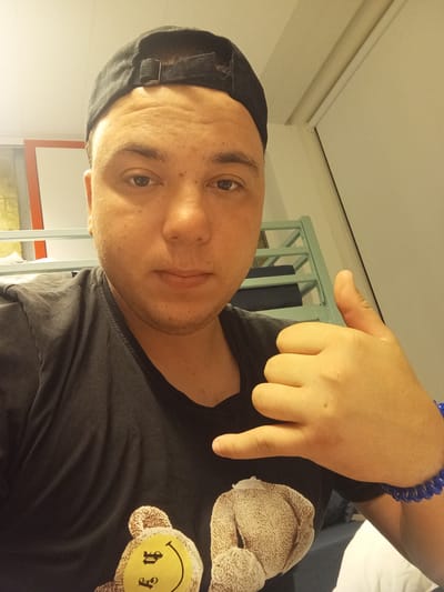 A person wearing a black baseball cap and a black t-shirt with a teddy bear graphic is shown in a close-up selfie. The person is making a hand gesture with their right hand, with their thumb and pinky extended. A blue beaded bracelet is visible on their wrist. The background appears to be an indoor setting, possibly a room with bunk beds, suggesting a hostel or dormitory. The lighting is warm and somewhat dim. The image is taken from a slightly high angle, looking down at the person. There is no identifiable information about the location in Anderlecht, Belgium.