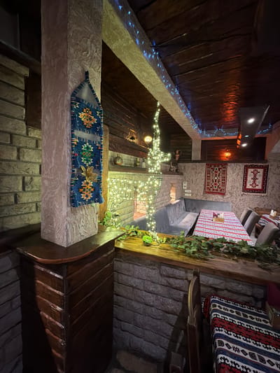 The image shows the interior of a restaurant in Kumbor, Montenegro. The decor features a rustic, ethnic style with stone and wood elements. A textured pillar is adorned with a woven tapestry. In the background, a fireplace is decorated with string lights, and two traditional rugs hang on the wall above a long, cushioned bench. A table with a striped tablecloth is set for dining, with chairs arranged around it. Green foliage trails along the edge of the wooden counter in the foreground. The ceiling is made of dark wooden planks and is decorated with blue string lights.