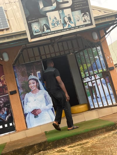 The image depicts the exterior of a photography studio located at NO. 4, AREWA PLAZA SABURI 1 DEI DEI, ABUJA, Nigeria. A man in a black t-shirt and black pants walks away from the camera towards an open metal gate. The studio features a large poster of a bride in a white wedding dress with a crown, positioned on the left side of the entrance. Above the entrance, a billboard advertises "AVID" photography services, listing "VIDEOS," "TRAININGS," "EVENTS," and "DRONE SHOTS." The billboard also displays a call line number: 07031685474. Adjacent to the bride's poster is another poster featuring a woman in a blue top holding a book. The studio is situated on a slightly elevated level, with a green mat extending from the entrance onto the ground. The overall environment suggests an outdoor commercial setting.