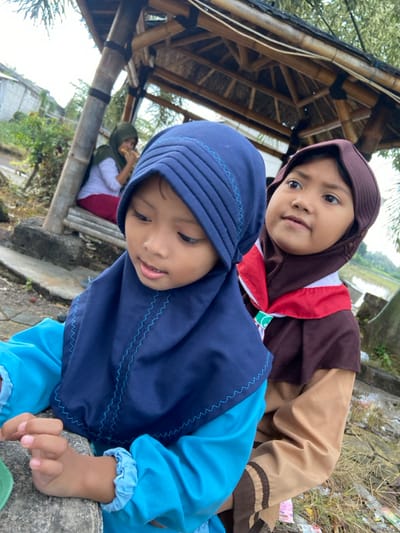 Three individuals are present outdoors in Kalitengah, Indonesia. In the foreground, two young girls are depicted. The girl on the left wears a dark blue hijab with light blue stitching and a light blue long-sleeved top. Her hands rest on a grey concrete surface, and she is looking downwards and slightly to her left. The girl on the right, partially obscured by the first, wears a brown hijab and a brown and tan uniform with a red and white neckerchief secured by a green woggle, indicative of a scout-like attire. She is looking upwards and to her right with a faint smile. In the mid-ground, a woman with an olive green hijab, white long-sleeved shirt, and red pants is seated on a bamboo bench within a bamboo-framed open-sided shelter or gazebo. She is engaged in eating an item held in her hands. The background reveals additional residential structures with concrete walls on the left, sparse vegetation, and a body of water, possibly a pond or flooded field, on the far right. The ground in the foreground is uneven, with dirt, gravel, and some dry grass.