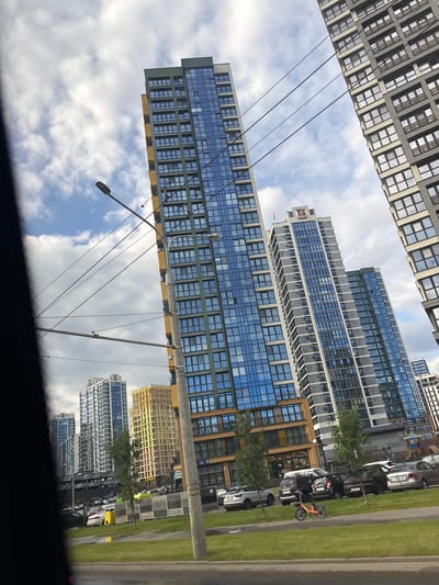 The image depicts a street-level view of a modern urban environment, likely in the village of Senitsky, Belarus, characterized by high-rise apartment buildings. The buildings are a prominent feature, showcasing contemporary architecture with large glass windows and varying facade designs, including a notable blue-tinted skyscraper and a yellow-hued building. Several cars are parked along the roadside, with a distinct orange electric scooter positioned on the grassy verge in the foreground, suggesting recent activity. Overhead power lines crisscross the sky, indicating the presence of public transportation or utility infrastructure. The sky is partly cloudy, with a mix of white clouds and patches of blue, suggesting a daytime scene. The overall impression is one of a developed residential area with a focus on modern design and infrastructure.